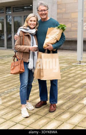 Una coppia senior in piedi sulla piazza lastricata fuori dall'entrata moderna tenendo borse della spesa e caffè Foto Stock
