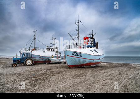 Tre barche da pesca sono adagiate su una spiaggia sabbiosa accanto a un trattore sotto un cielo coperto, evocando l'industria costiera, la solitudine tranquilla e il tra marittimo Foto Stock