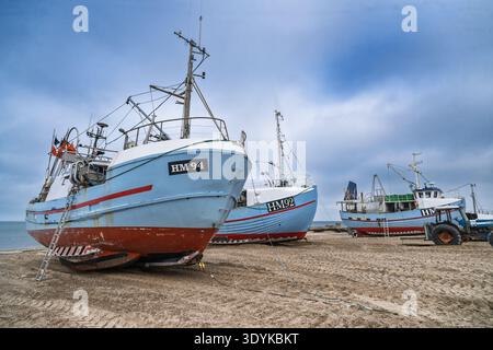 Tre barche da pesca sono adagiate su una spiaggia sabbiosa accanto a un trattore sotto un cielo coperto, evocando l'industria costiera, la solitudine tranquilla e il tra marittimo Foto Stock