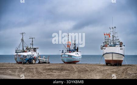 Tre barche da pesca sono adagiate su una spiaggia sabbiosa accanto a un trattore sotto un cielo coperto, evocando l'industria costiera, la solitudine tranquilla e il tra marittimo Foto Stock