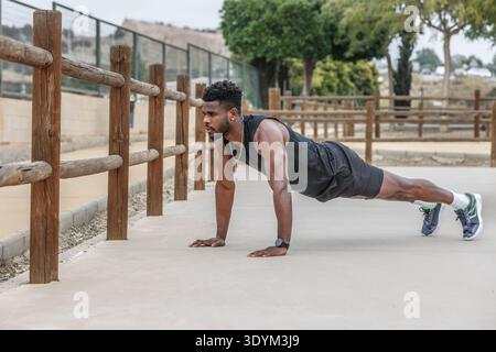 Corpo completo di un forte atleta afroamericano che esegue un esercizio su una tavola a terra vicino a una recinzione di legno durante l'allenamento fisico Foto Stock
