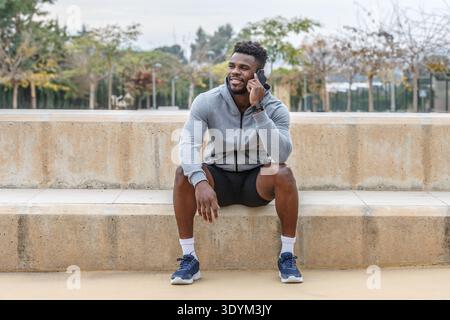 Corpo pieno di giovane atleta afroamericano felice in abbigliamento sportivo seduto sulle scale e a parlare tramite telefono cellulare durante la pausa dall'allenamento Foto Stock