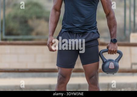 Taglia un atleta nero anonimo maschile con un abbigliamento sportivo stretto che regge il kettlebell pesante mentre ti alleni sul campo sportivo in città Foto Stock