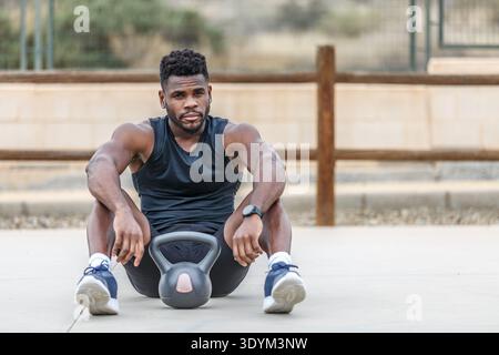 Giovane sportivo con barba etnica e muscoloso in abbigliamento sportivo e orologio intelligente seduto sul pavimento con kettlebell durante l'allenamento all'aperto mentre guardi Foto Stock