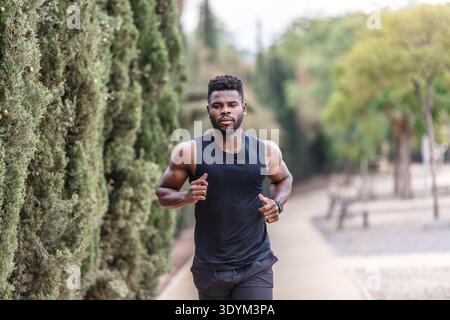 Uomo nero sicuro di sé con barba e muscoloso che fa jogging lungo il parco durante l'allenamento cardio-fitness il giorno d'estate contro gli alberi sfocati Foto Stock