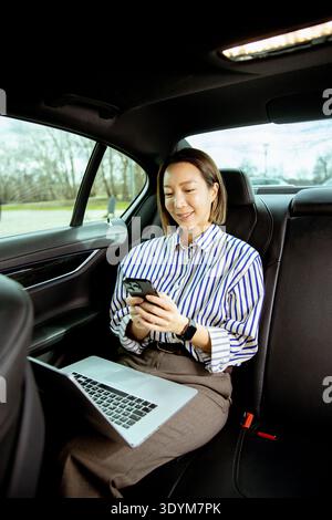 Una donna coreana siede sul sedile posteriore di un'auto. Sorride mentre usa il suo smartphone. Un notebook si appoggia sulle sue ginocchia mentre si mette in contatto con i suoi dispositivi. TH Foto Stock
