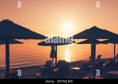 Ombrelloni di paglia e lettini vuoti si stagliano contro il sole nascente sulla spiaggia di Policrono a Halkidiki, in Grecia. Il mare tranquillo riflette la sua bellezza Foto Stock
