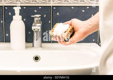 Focalizzazione selettiva sulla mano tenendo la spugna schiumosa accanto alla bottiglia di sapone e sul lavello in porcellana. Foto Stock
