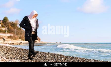 Un giovane maschio con cappotto incappucciato si erge in modo contemplativo sulla spiaggia di ciottoli con onde che si infrangono e orizzonte chiaro. Foto Stock