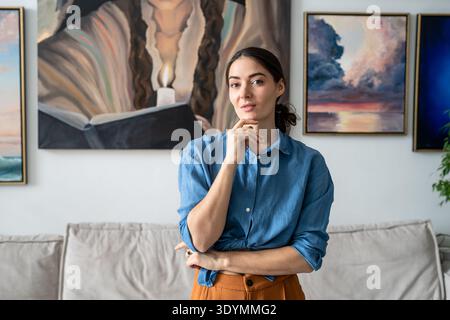 Una donna premurosa che posa alla macchina fotografica nella galleria del loft di casa con dipinti installati sulla parete. Foto Stock