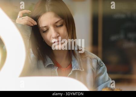 La giovane bruna in giacca di denim ha un aspetto penoso e leggermente triste mentre si riposa la testa a mano in un accogliente ambiente da caffè con una calda illuminazione Foto Stock