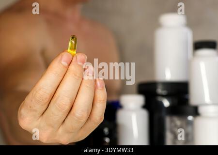 Primo piano di una capsula di olio di pesce dorato con bottiglie di supplemento sullo sfondo. Uomo muscolare che tiene in finge la capsula softgel dell'olio di pesce omega-3 Foto Stock