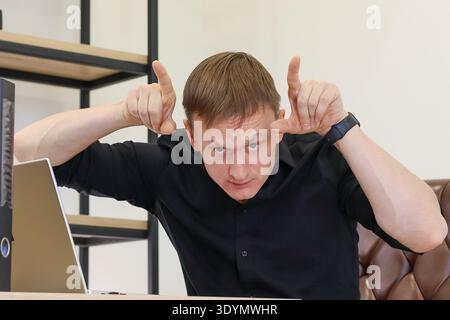 Ha agitato il giovane dirigente nell'abbigliamento formale formando corna del diavolo con le mani alzate alla scrivania dell'ufficio vicino allo schermo del computer. Foto Stock