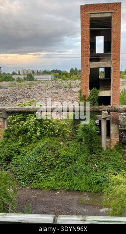 Un'alta struttura in mattoni si erge circondata da fitte piante verdi e detriti. L'area mostra segni di abbandono e abbandono con cemento rotto e sca Foto Stock
