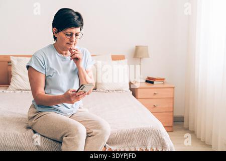 Donna caucasica matura seduta sul letto con smartphone in una camera da letto accogliente Foto Stock