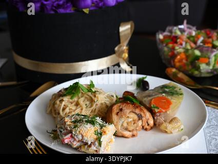Spaghetti alla carbonara, pollo, aspico e olive su un piatto bianco con rose blu sullo sfondo Foto Stock