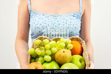 Donna in abito blu che tiene in mano un cestino di vimini pieno di frutta biologica fresca. Raccolta estiva, nutrizione sana e concetto di stile di vita vegano Foto Stock