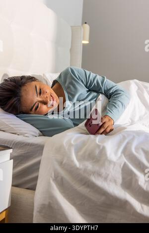 Donna asiatica sdraiata sul letto che indossa una parte superiore blu a maniche lunghe che regge lo smartphone rosa e sorride Foto Stock