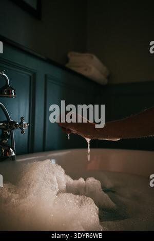 Le mani scaricano l'acqua dal bagno riempito di bolle in un bagno soffice e soffice. L'illuminazione soffusa mette in risalto il vapore e la consistenza della schiuma. Moma sereno, rilassante e intimo Foto Stock