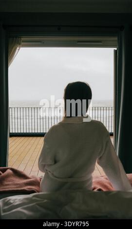 La persona siede sul letto e guarda il mare dal balcone. Grigio cielo, calma, tranquilla. Illuminazione soffusa, atmosfera accogliente. Ideale per viaggi, benessere o relax Foto Stock