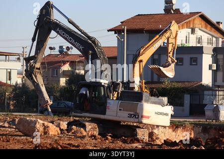 Escavatore cingolato idraulico per impieghi gravosi che lavora in un cantiere polveroso con dumper ed edifici residenziali sullo sfondo Foto Stock