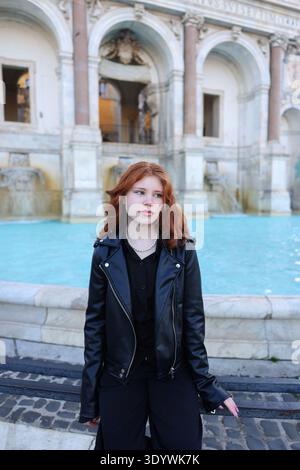Ritratto di una giovane rossa seduta accanto a una fontana storica con architettura classica in una città europea. Foto Stock