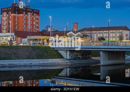 VINKOVCI, CROAZIA - 18 febbraio 2026: Tranquilla vista serale degli edifici e degli alberi che si riflettono in un fiume calmo, illuminato da calde luci stradali Foto Stock