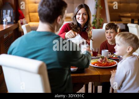 Una famiglia condivide la colazione ad un tavolo in un caffè. La madre sorride mentre il padre versa il latte. Due bambini sembrano felici mentre si siedono con cibo colorato Foto Stock