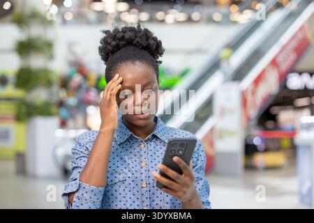 Una giovane donna che si sente turbata e stressata mentre riceve notizie preoccupanti sul suo cellulare, soffre di mal di testa e preoccupazione all'interno di un vivace centro commerciale moderno Foto Stock