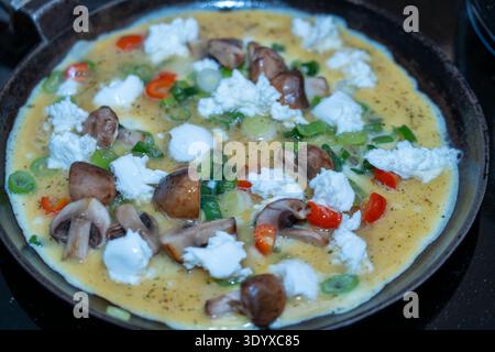 Primo piano con angolo superiore di una frittata all'uovo ricca di proteine, ricoperta da formaggio bianco, funghi e peperoni, che cuoce su una stufa a induzione. Foto Stock