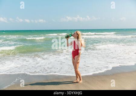 Giovane donna in costume da bagno rosso che tiene in mano una bottiglia d'acqua a Sunny Beach Foto Stock