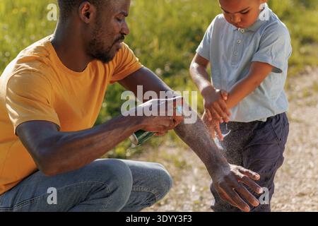 Padre spruzza repellente per insetti sul bambino in un'area verde all'aperto mentre si prepara per un'escursione per evitare morsi da insetti. Foto Stock