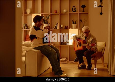 Senior Black Woman che fotografa Senior Caucasian Man che suona la chitarra al chiuso Foto Stock