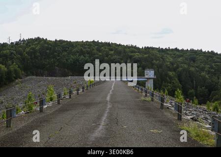 Strada vuota che attraversa una grande diga circondata da colline boscose e alberi verdi in un paesaggio naturale. Foto Stock