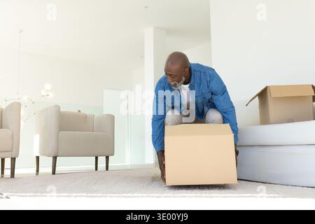 Uomo afroamericano senior che maneggia una scatola mobile su un tappeto nel moderno soggiorno con giacca in denim. Anziani, divano, poltrone, interni, disimballaggio, sole Foto Stock