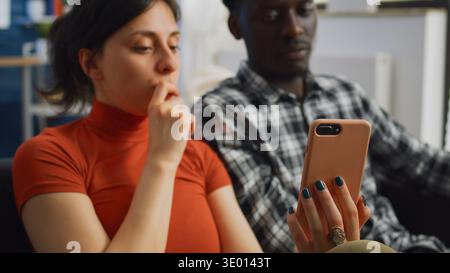 Primo piano di coppia interrazziale guardando lo schermo dello smartphone parlare mentre si siede sul divano in salotto. Uomo e donna multi etnico sposato utilizzando la tecnologia su gadget moderno a casa Foto Stock