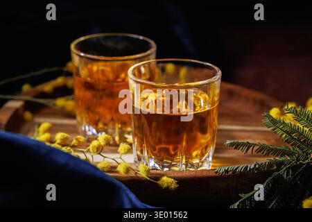 Due bicchieri di whisky ambrato con fiori di mimosa su vassoio in legno, celebrazione primaverile Foto Stock