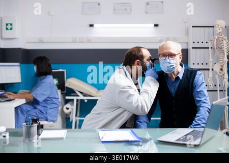 Il giovane medico usa l'otoscopio su un anziano malato seduto in clinica, garantendo una diagnosi accurata durante la consultazione sanitaria. Otologo maschio che fa l'esame dell'orecchio su un paziente pensionato con strumento medico. Foto Stock