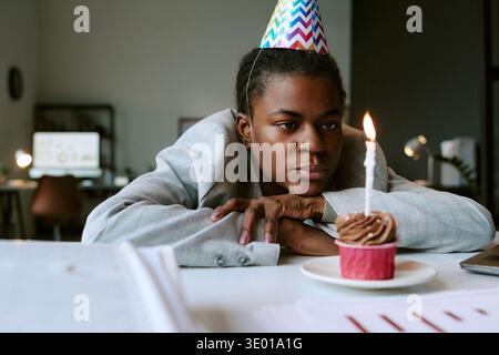 Giovane donna nera adulta che indossa un cappello da festa che fissa la candela per la torta di compleanno Foto Stock
