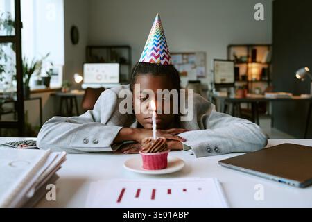 Giovane donna nera adulta che indossa il cappello da festa che fissa la torta di compleanno illuminata Foto Stock