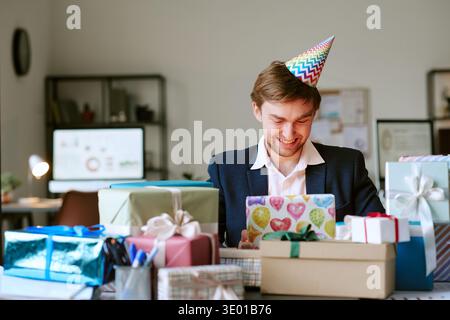 Giovane uomo caucasico che indossa il cappello da festa sorridente mentre apre i regali di compleanno Foto Stock