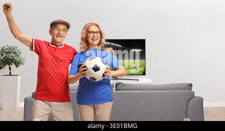 Una coppia di anziani sorridente che tiene una partita di calcio e tifa davanti a una tv Foto Stock
