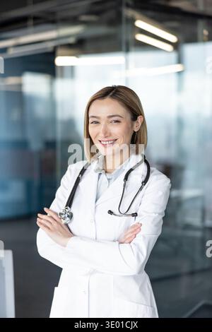 Medico donna in piedi con le braccia incrociate, sorridente alla telecamera, che comunica professionalità medica, competenza e fiducia in un ambiente sanitario moderno Foto Stock