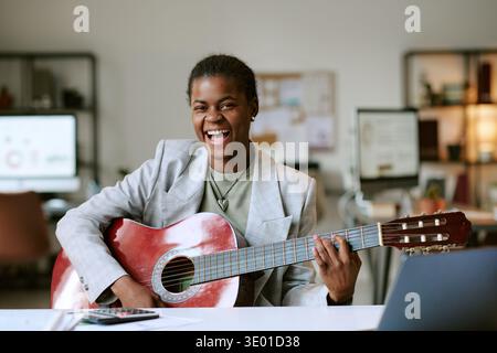 Ritratto della giovane donna nera adulta sorridente mentre suona la chitarra acustica in interni Foto Stock