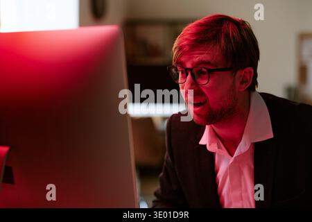 Giovane uomo caucasico che indossa occhiali che reagiscono allo schermo del computer in interni Foto Stock