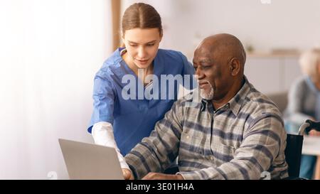 L'uomo anziano impara a utilizzare un notebook con l'aiuto di una giovane donna in un ambiente di cura Foto Stock
