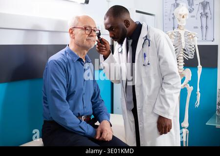 Specialista della ent afroamericana che fa l'esame dell'orecchio su un uomo anziano seduto sul letto d'ospedale in ufficio della clinica. Esperto medico nero che tiene l'otoscopio verso il paziente anziano durante il controllo medico. Foto Stock