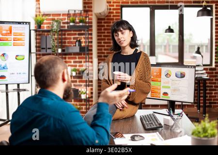 Donna manager seduto al tavolo e tenendo in mano una tazza di caffè mentre la dipendente spiega la strategia di marketing per l'azienda startup. Donna d'affari che ascolta il giovane uomo che descrive l'analisi finanziaria in un ufficio a muro. Foto Stock