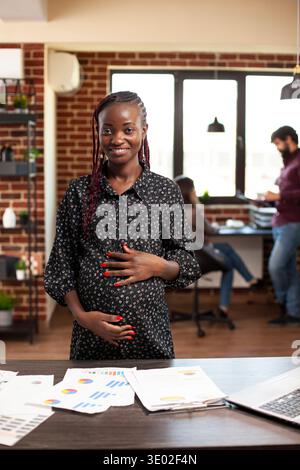 Ritratto di una donna d'affari di colore incinta in piedi accanto alla scrivania piena di documenti di ricerca. Sorridente madre in attesa afroamericana con le mani sulla pancia della gravidanza e guardando la macchina fotografica. Foto Stock