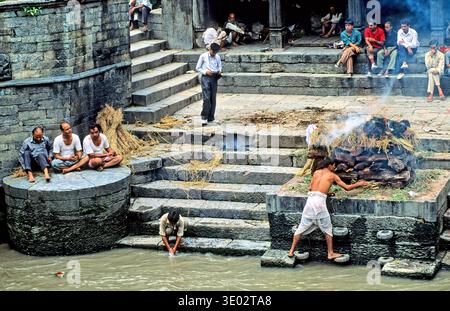 Le persone osservano una cerimonia di cremazione a Pashupatinath a Kathmandu, mentre altri si siedono nelle vicinanze e osservano il flusso del fiume. Foto Stock